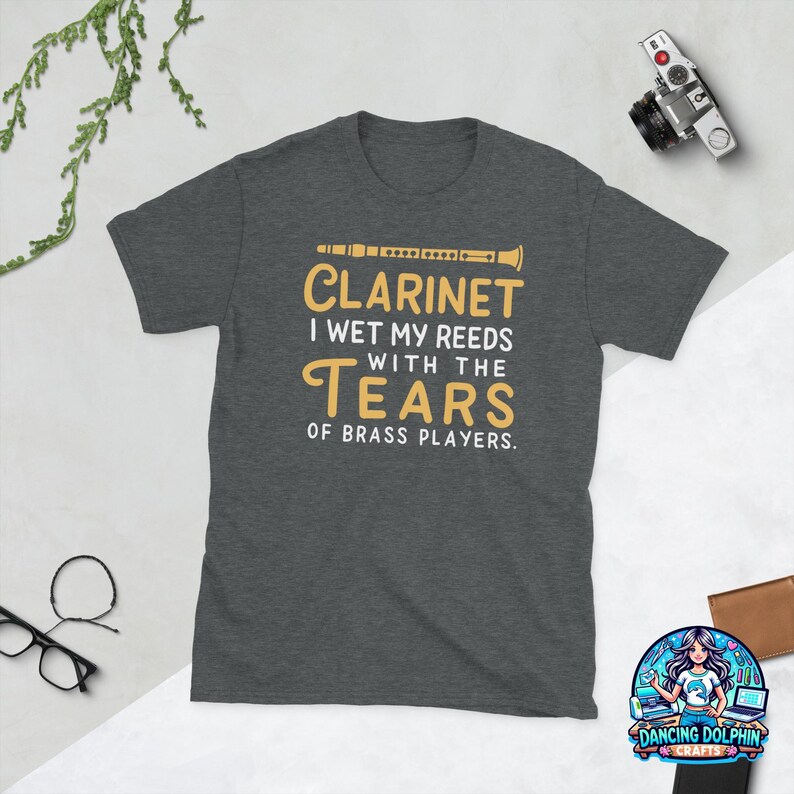 May include: A dark heather gray short-sleeve t-shirt featuring a graphic of a clarinet and the text: CLARINET I WET MY REEDS WITH THE TEARS OF BRASS PLAYERS. The design uses yellow and white lettering centered on the chest of the garment.