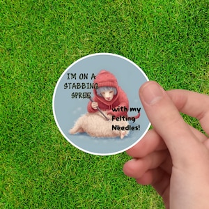 May include: A round sticker with a cartoon illustration of a person in a red hoodie stabbing a fluffy white sheep with a felting needle. The text on the sticker reads "I'm on a stabbing spree with my felting needles!"