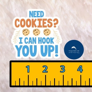 May include: A white sticker with the text "NEED COOKIES? I CAN HOOK YOU UP!" and three chocolate chip cookies. The sticker is on a white fuzzy surface with a ruler underneath. The ruler is yellow with black markings and has the numbers 1, 2, 3, and 4.
