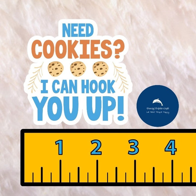 May include: A white sticker with the text "NEED COOKIES? I CAN HOOK YOU UP!" and three chocolate chip cookies. The sticker is on a white fuzzy surface with a ruler underneath. The ruler is yellow with black markings and has the numbers 1, 2, 3, and 4.