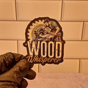 May include: A brown and black sticker with a cartoon illustration of a person using a circular saw to cut wood. The text "WOOD Whisperer" is written in white letters on the sticker.