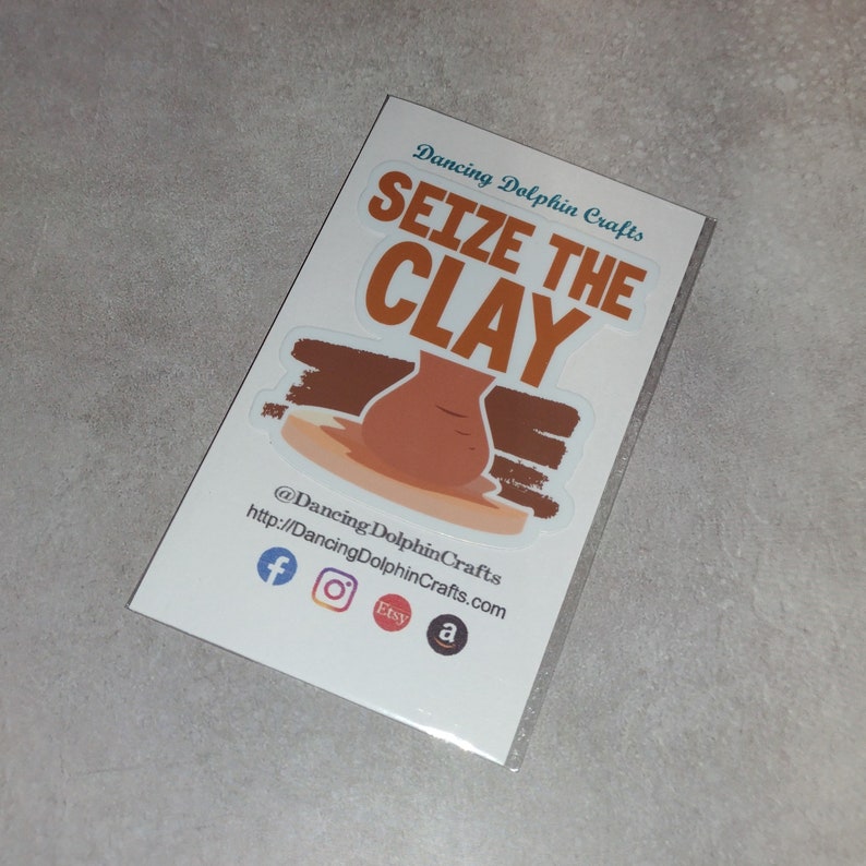 K&ouml;nnte beinhalten: Ein Aufkleber mit einem braunen Tontopf auf einer T&ouml;pferscheibe und dem Text "Seize the Clay" in Orange. Der Aufkleber stammt von Dancing Dolphin Crafts und enth&auml;lt deren Social-Media-Handles und Website.