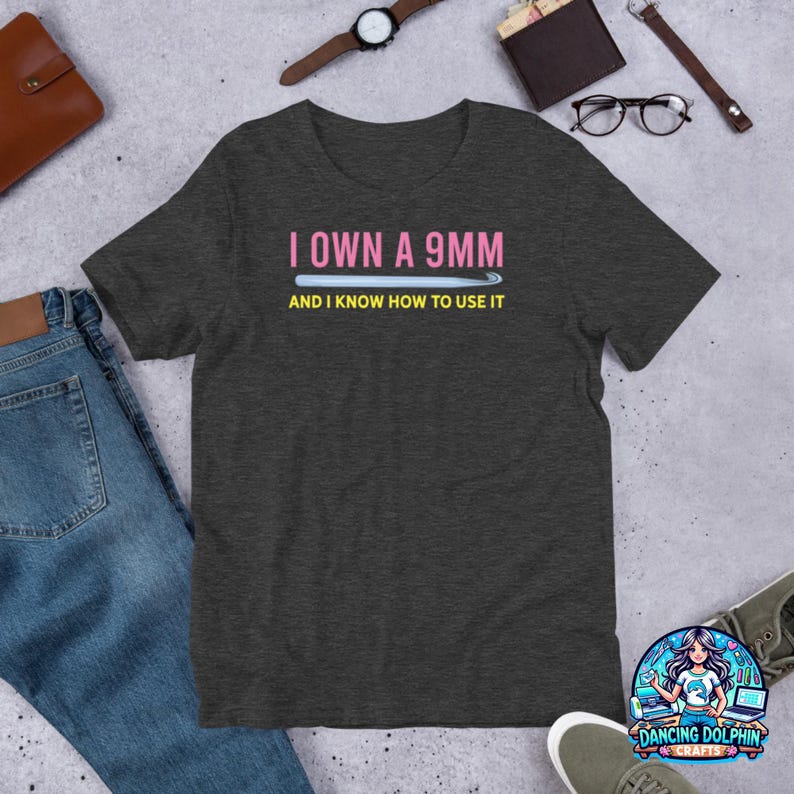 K&ouml;nnte beinhalten: Dunkelgraues T-Shirt mit einem pinkfarbenen und wei&szlig;en Aufdruck mit der Aufschrift "I own a 9mm and I know how to use it".