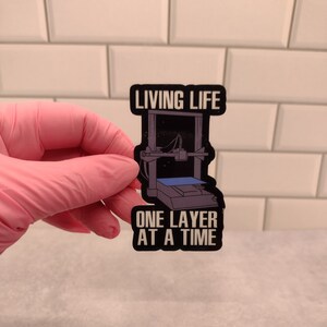 May include: A black sticker featuring a 3D printer illustration with the text "LIVING LIFE" above and "ONE LAYER AT A TIME" below. The sticker is held by a pink gloved hand against a white tiled background.