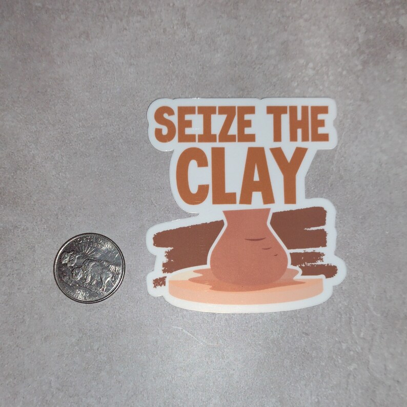 K&ouml;nnte beinhalten: Ein wei&szlig;er Aufkleber mit dem Text "Seize the Clay" in braunen Buchstaben. Ein brauner Tontopf steht auf einer T&ouml;pferscheibe, mit braunen Pinselstrichen dahinter.