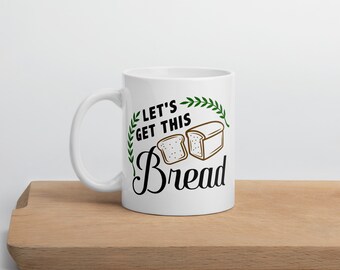 Könnte beinhalten: Weiße Keramik-Tasse mit dem Text "LET'S GET THIS Bread" in schwarzer Schrift und einer Brotlaib-Illustration. Die Tasse hat ein grünes Blattdesign um den Text und die Illustration. Die Tasse steht auf einer Holzoberfläche.