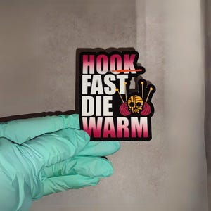 May include: A pink and black sticker with the text "HOOK FAST DIE WARM" and a cartoon image of a skull wearing a pink and red yarn ball.