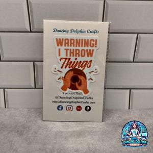 May include: A white sticker with the text "WARNING! I THROW Things" in orange and brown. The sticker features a graphic of hands throwing a brown object. The sticker also includes social media icons and the text "Dancing Dolphin Crafts".