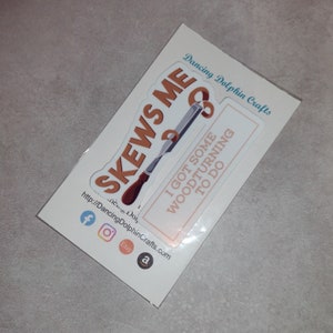 May include: A white sticker with a brown woodturning skew chisel and the text "SKEWS ME" and "I GOT SOME WOODTURNING TO DO". The sticker is on a white background with the text "Dancing Dolphin Crafts" and a website address.