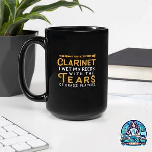 May include: A black ceramic mug featuring a yellow graphic of a clarinet and the text: CLARINET I WET MY REEDS WITH THE TEARS OF BRASS PLAYERS. The mug has a curved handle and a smooth, glossy finish, displaying clear, bold lettering.