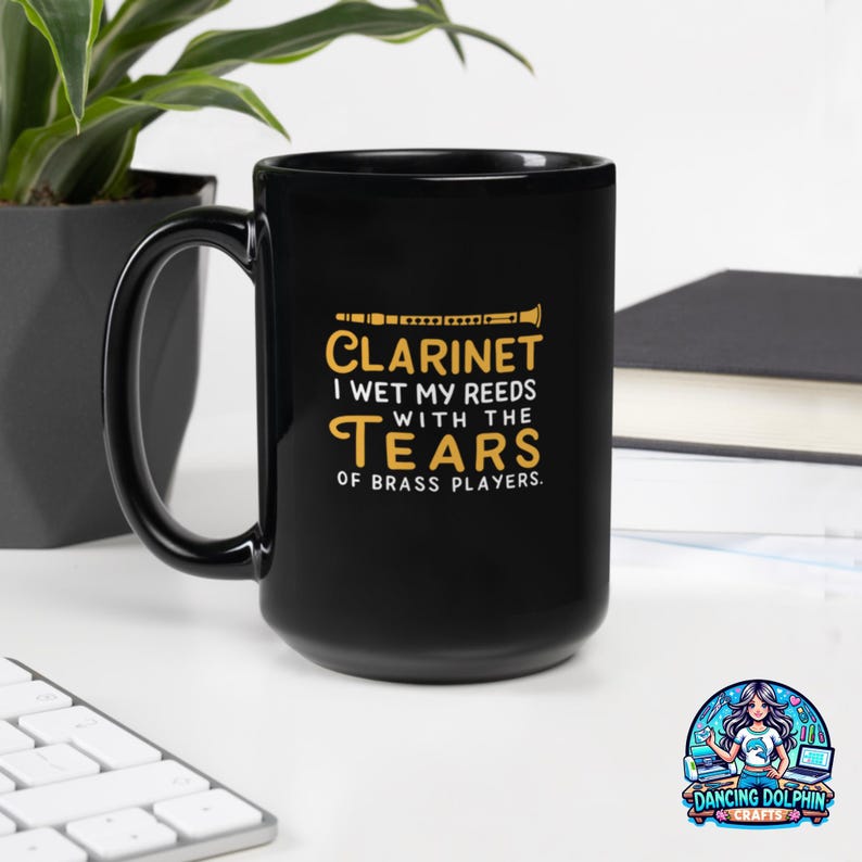 May include: A black ceramic mug featuring a yellow graphic of a clarinet and the text: CLARINET I WET MY REEDS WITH THE TEARS OF BRASS PLAYERS. The mug has a curved handle and a smooth, glossy finish, displaying clear, bold lettering.