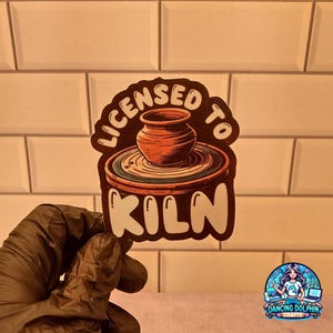 May include: A sticker with the text "LICENSED TO KILN" in white, arched over a pottery wheel with a brown pot. The sticker has a black border and is held by a gloved hand. The background is a white tiled wall.