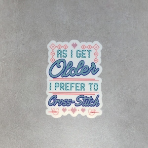 May include: White sticker with a blue and red cross-stitch design. The text reads "As I get older I prefer to cross-stitch."