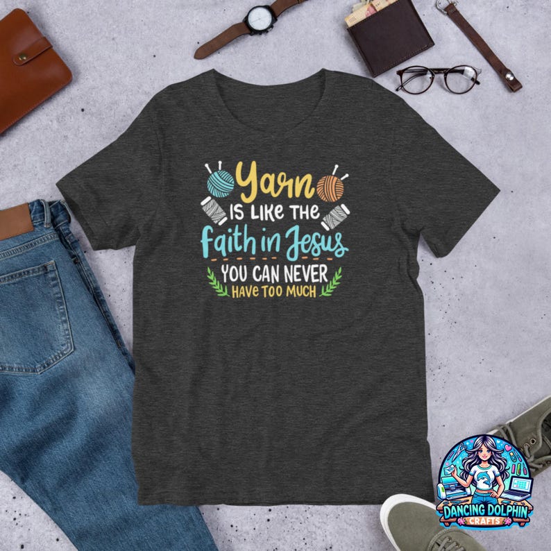 May include: Dark gray t-shirt with the text "Yarn is like the faith in Jesus, you can never have too much." The shirt features illustrations of yarn balls in blue and orange. A fun tee for knitters and crocheters.