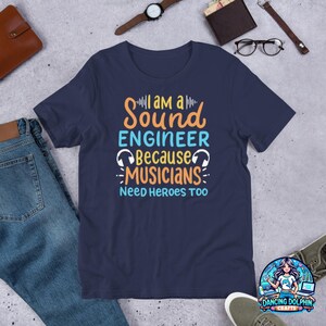 以下が含まれることがあります： 紺色のTシャツに、「私はサウンドエンジニアです。なぜならミュージシャンはヒーローを必要とするからです」というグラフィックがプリントされています。グラフィックには、スタイリッシュなサウンドウェーブとヘッドホンが描かれています。