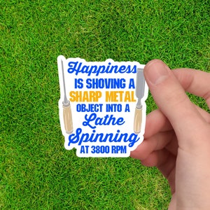 May include: A blue and yellow sticker with the text "Happiness is shoving a sharp metal object into a lathe spinning at 3800 RPM". The sticker features two wood chisels.