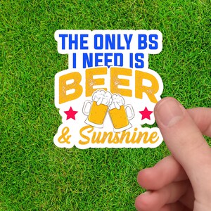 Könnte beinhalten: Weißer Aufkleber mit dem Text "The only BS I need is Beer & Sunshine" in Blau und Gelb. Zwei Cartoon-Bierkrüge stoßen an, mit roten Sternen auf beiden Seiten.