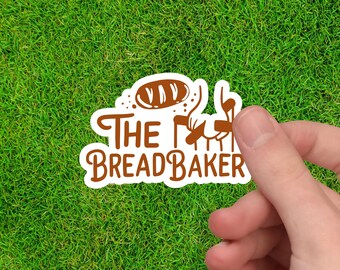 May include: A white sticker with brown text and illustrations, held against a backdrop of green grass. The sticker reads "The BreadBaker" with a bread loaf illustration above the text. The design is a playful, food-themed graphic.