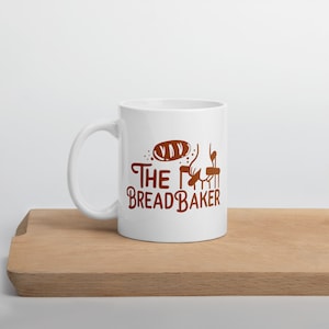 May include: White ceramic mug with a brown graphic that says "The Bread Baker" and a cartoon image of a loaf of bread and a hand holding a rolling pin.
