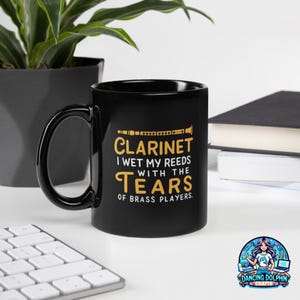 May include: A black ceramic mug featuring a yellow graphic of a clarinet and the text: CLARINET I WET MY REEDS WITH THE TEARS OF BRASS PLAYERS. The mug has a curved handle and a smooth, glossy finish, displaying clear, bold lettering on the front side.