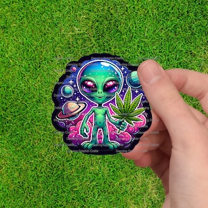 May include: A colorful sticker featuring a cartoon alien holding a cannabis leaf. The alien is green with large purple eyes, set against a backdrop of planets, stars, and pink clouds. The sticker has the text "Dancing Dolphin Crafts".