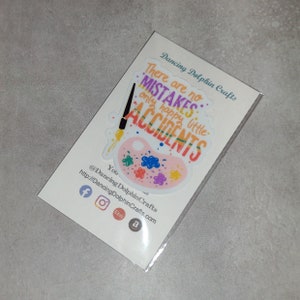 Puede incluir: Una pegatina con una paleta de pintura rosa y un pincel con el texto "No hay errores, solo peque&ntilde;os accidentes felices." La pegatina est&aacute; en una tarjeta blanca con el texto "Dancing Dolphin Crafts" y iconos de redes sociales.