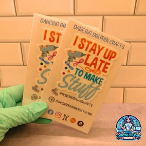 May include: Two stickers with a light blue background and colorful text. The text on the stickers reads "I stay up late to make stuff". The stickers are on a white background with a logo for Dancing Dolphin Crafts.