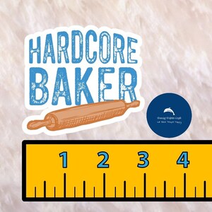 May include: A white sticker with blue text that reads "HARDCORE BAKER" and a brown rolling pin. A blue circle sticker with a dolphin graphic and the text "Sharing Dolphin Joy! We Make Space Happy" is in the background.