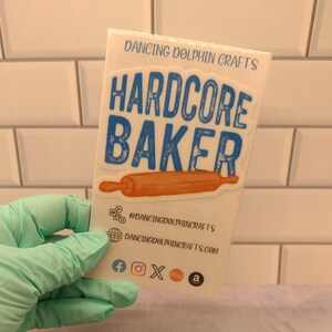 May include: A white card with blue and orange text that reads "Hardcore Baker" with a rolling pin graphic. The card also includes the text "Dancing Dolphin Crafts" and social media icons for Facebook, Instagram, Etsy, and Amazon.