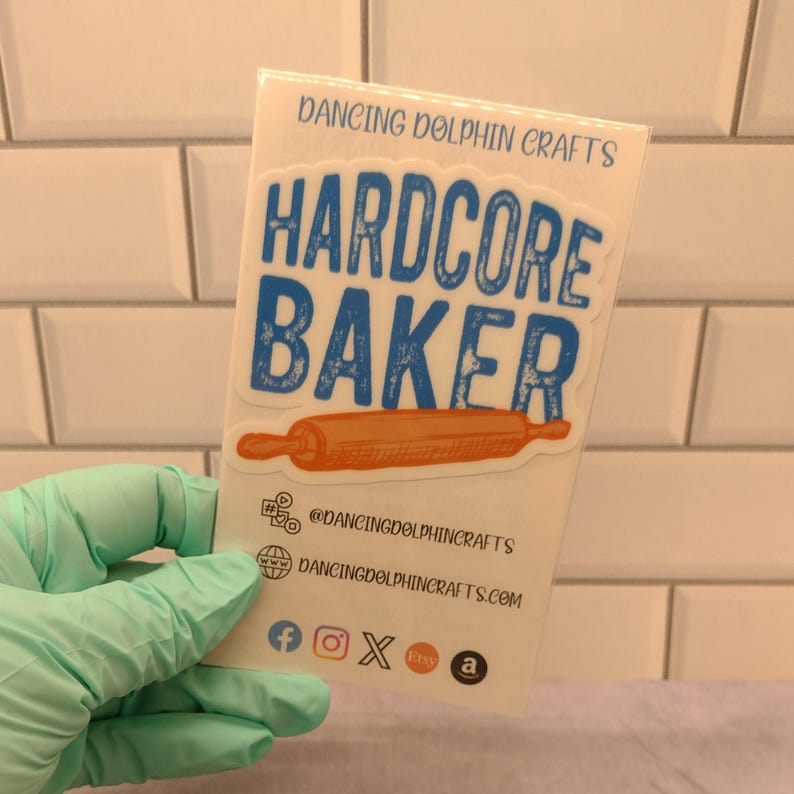 May include: A white card with blue and orange text that reads "Hardcore Baker" with a rolling pin graphic. The card also includes the text "Dancing Dolphin Crafts" and social media icons for Facebook, Instagram, Etsy, and Amazon.