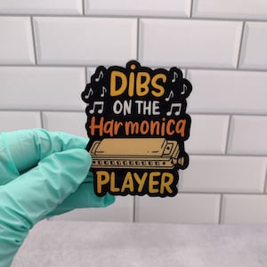 May include: Black and yellow sticker with the text "Dibs on the Harmonica Player" and a cartoon harmonica.