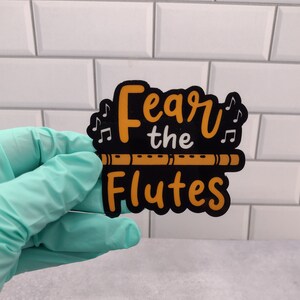 May include: A black and orange sticker with the text "Fear the Flutes" and a small brown flute graphic. The sticker is held in a gloved hand.