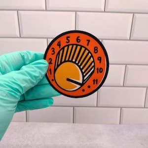 May include: Orange and black circular sticker with a graphic of a dial with numbers 1 through 11. The dial is set to 5.