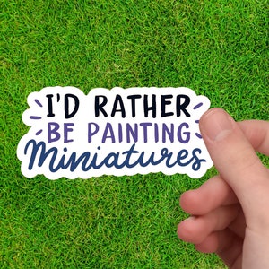 May include: A white sticker with the text "I'd rather be painting miniatures" in black, purple, and blue. The sticker is held by a hand against a green grass background.