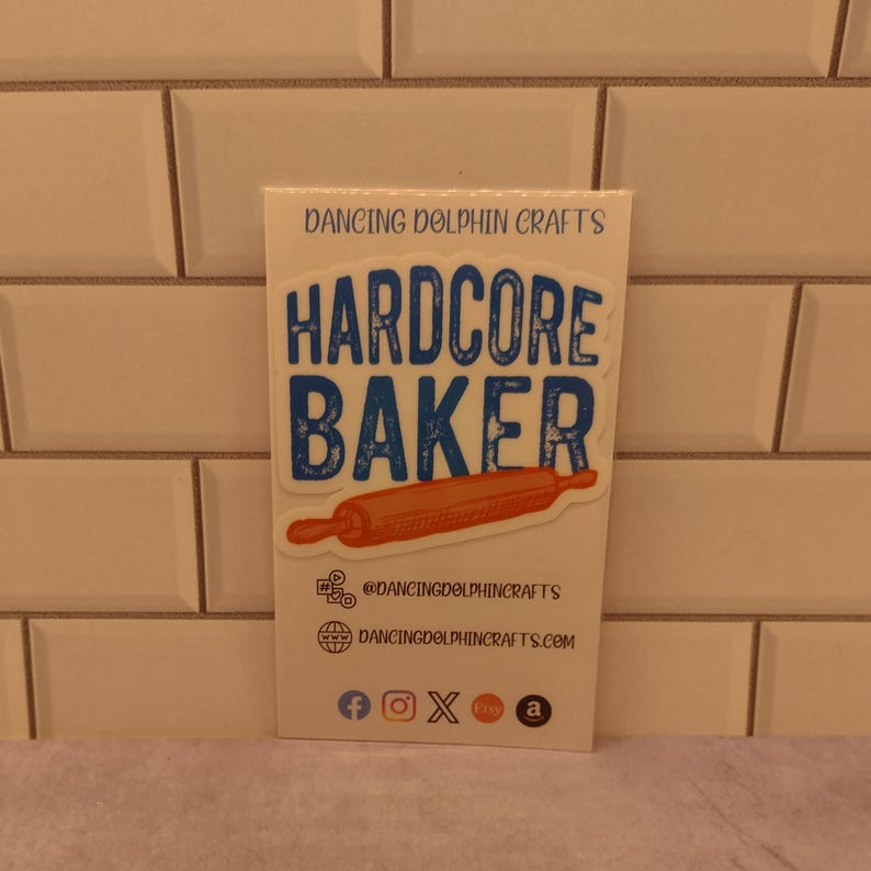 May include: A white sticker with blue and orange text that reads "HARDCORE BAKER" with a rolling pin graphic. The sticker is on a white background with the text "DANCING DOLPHIN CRAFTS" at the top and social media icons at the bottom.
