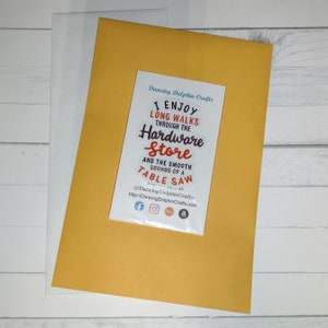 May include: A yellow card with a white sticker that says "I enjoy long walks through the hardware store and the smooth sounds of a table saw." The sticker also includes the text "Dancing Dolphin Crafts" and social media icons for Facebook, Instagram, Etsy, and Amazon.