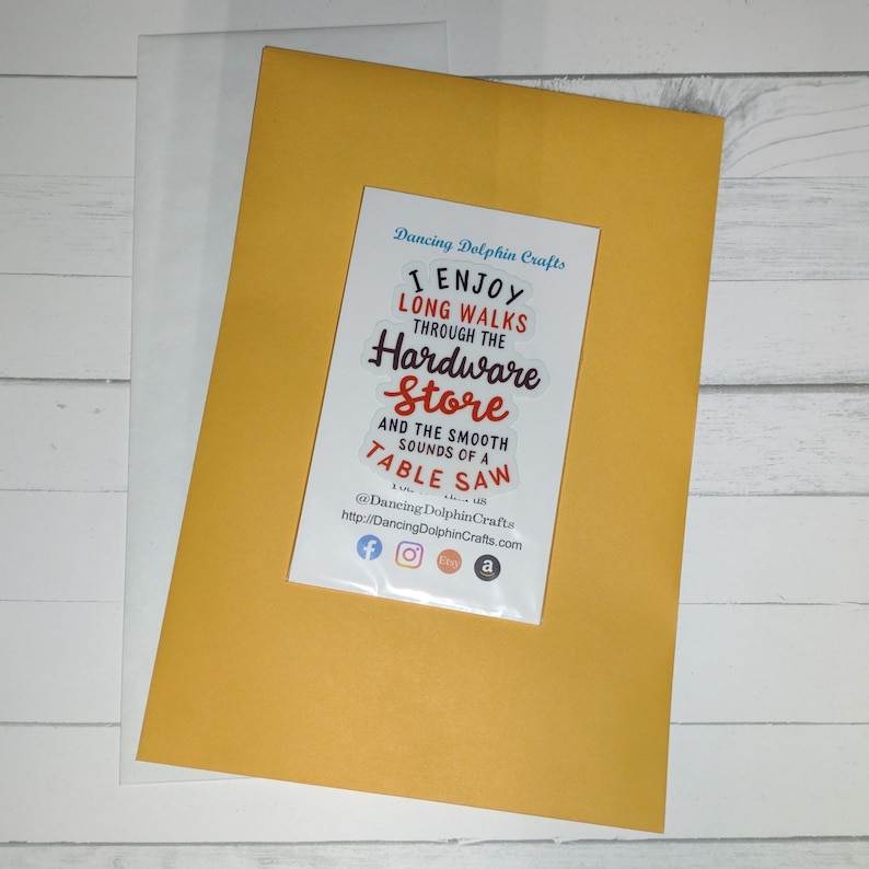 May include: A yellow card with a white sticker that says "I enjoy long walks through the hardware store and the smooth sounds of a table saw." The sticker also includes the text "Dancing Dolphin Crafts" and social media icons for Facebook, Instagram, Etsy, and Amazon.