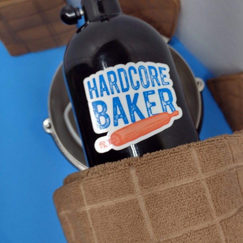 May include: A black bottle with a sticker that says "HARDCORE BAKER" and a red rolling pin illustration. The bottle is sitting on a brown surface.