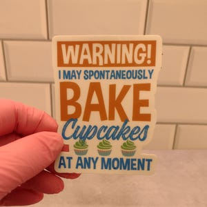 May include: White sticker with the text "WARNING! I MAY SPONTANEOUSLY BAKE CUPCAKES AT ANY MOMENT". The text is in a blue and orange font. There are three green cupcakes with blue frosting and white sprinkles on the sticker.