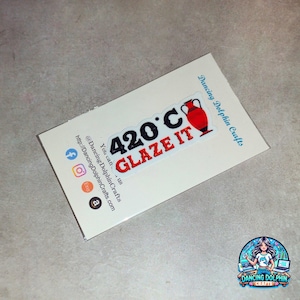 May include: A white sticker with black and red text that reads "420°C GLAZE IT". A red clay pot icon is next to the text. The sticker is on a white background with the text "Dancing Dolphin Crafts" and social media icons.