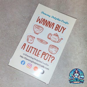 May include: A sticker sheet with the text "WANNA BUY A LITTLE POT?" in red, along with illustrations of teacups, a teapot, and a saucer. The sticker sheet also includes social media handles and a logo for Dancing Dolphin Crafts.