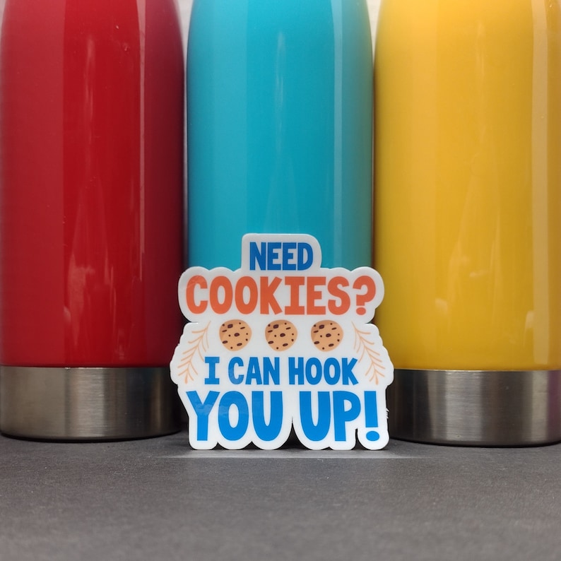 May include: A white sticker with blue, orange, and brown text and images. The sticker says "Need Cookies? I can hook you up!" and has three images of chocolate chip cookies.