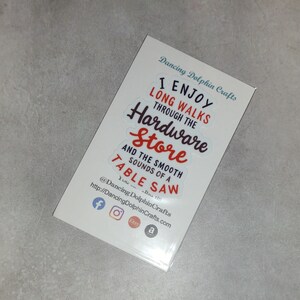 May include: A white card with the text "I enjoy long walks through the hardware store and the smooth sounds of a table saw" in red and black lettering. The card also includes social media icons for Facebook, Instagram, Etsy, and Amazon.
