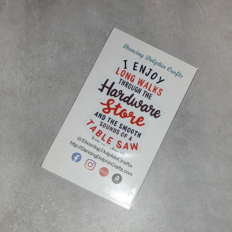 May include: A white card with the text "I enjoy long walks through the hardware store and the smooth sounds of a table saw" in red and black lettering. The card also includes social media icons for Facebook, Instagram, Etsy, and Amazon.
