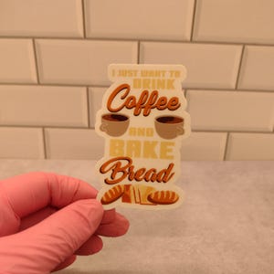 May include: White sticker with the text "I just want to drink coffee and bake bread". The text is in a brown font with a yellow outline. There are two coffee cups and three loaves of bread on the sticker.