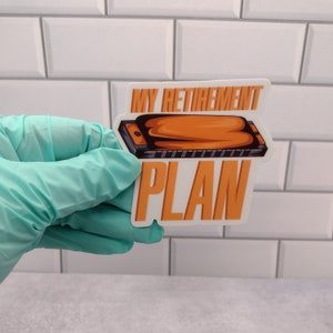 May include: A sticker with the text "MY RETIREMENT PLAN" in orange block letters. The image features an orange and black harmonica. The sticker is held by a person wearing a teal glove.