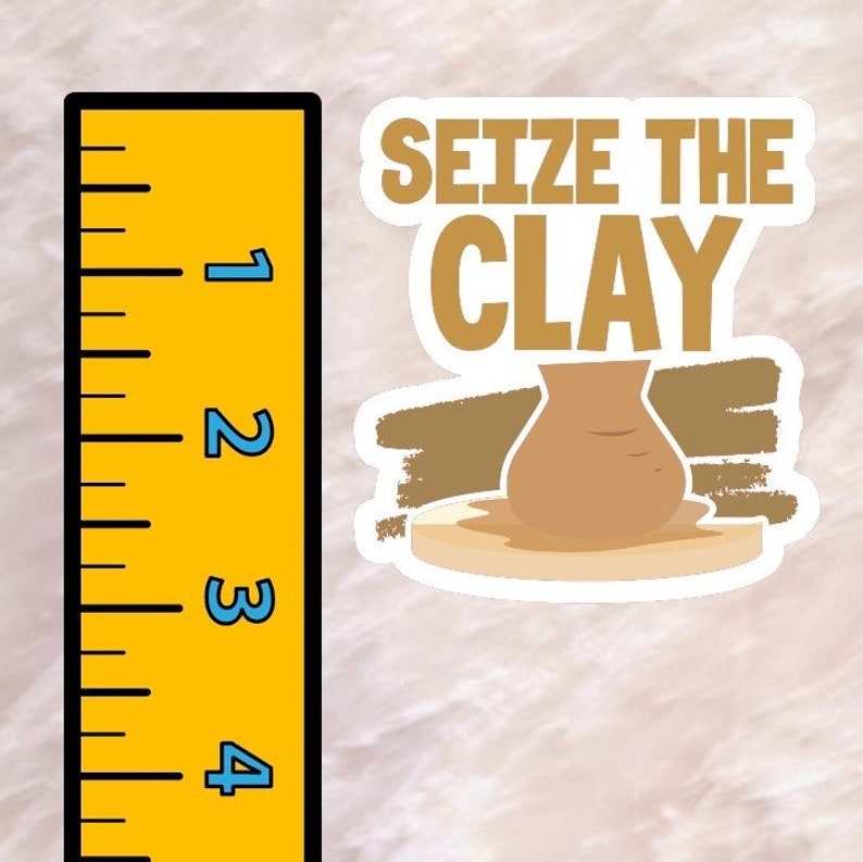 K&ouml;nnte beinhalten: Ein wei&szlig;er Aufkleber mit braunem und goldenem Text, der "Seize the Clay" lautet. Ein brauner Tontopf steht auf einer wei&szlig;en T&ouml;pferscheibe.