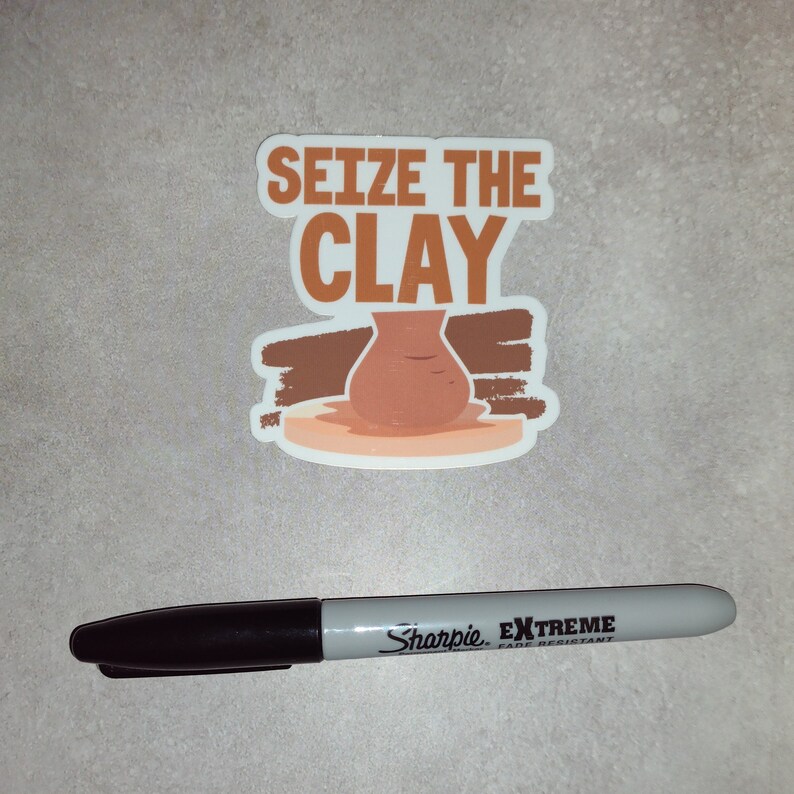 K&ouml;nnte beinhalten: Ein brauner und wei&szlig;er Aufkleber mit dem Text "Seize the Clay" und einem Cartoon-Bild eines Tontopfs auf einer T&ouml;pferscheibe.