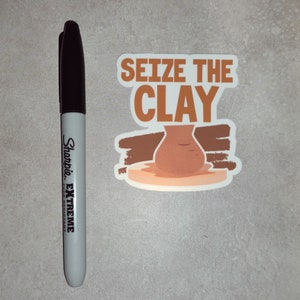 K&ouml;nnte beinhalten: Ein wei&szlig;er Aufkleber mit braunen und orangen Akzenten. Der Aufkleber zeigt eine Tonvase auf einer Plattform und den Text "Seize the Clay".