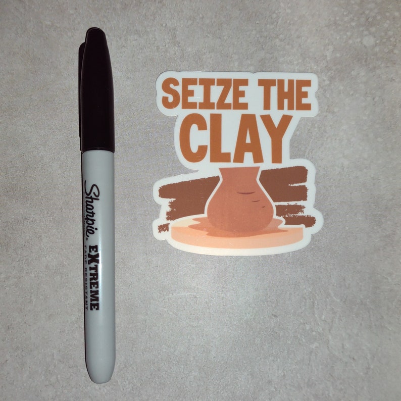 K&ouml;nnte beinhalten: Ein wei&szlig;er Aufkleber mit braunen und orangen Akzenten. Der Aufkleber zeigt eine Tonvase auf einer Plattform und den Text "Seize the Clay".